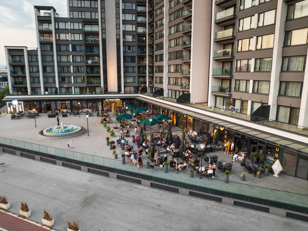 The square in front of River Walk at dusk, with the fountain and café tables below the apartments